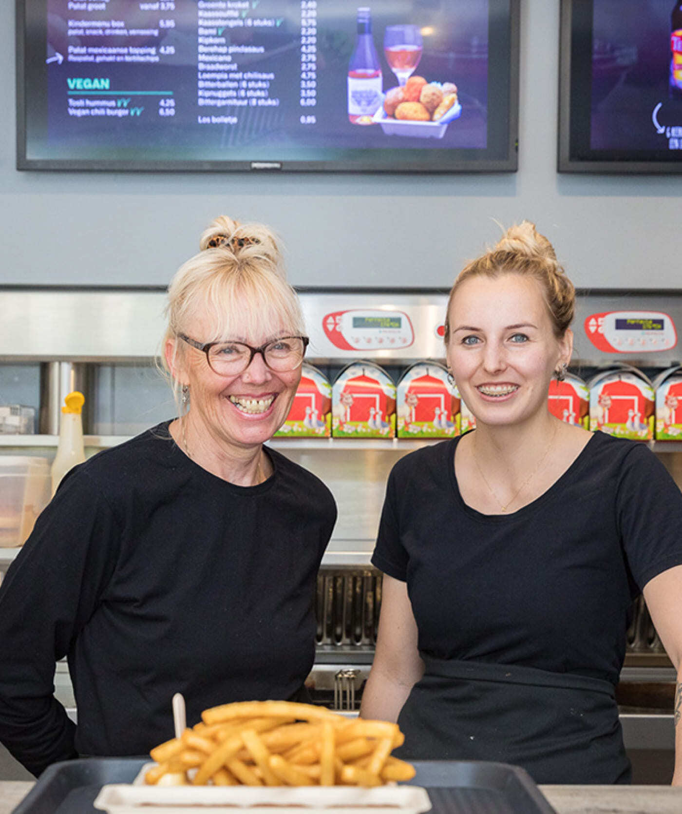 Twee vrouwen in zwarte shirts staan achter een toonbank met een schaal frieten. Ze lachen, met op de achtergrond de menukaarten en de sauzen zichtbaar. Ze staan klaar om klanten te bedienen of om hun cateringwebsites en online diensten te bespreken.