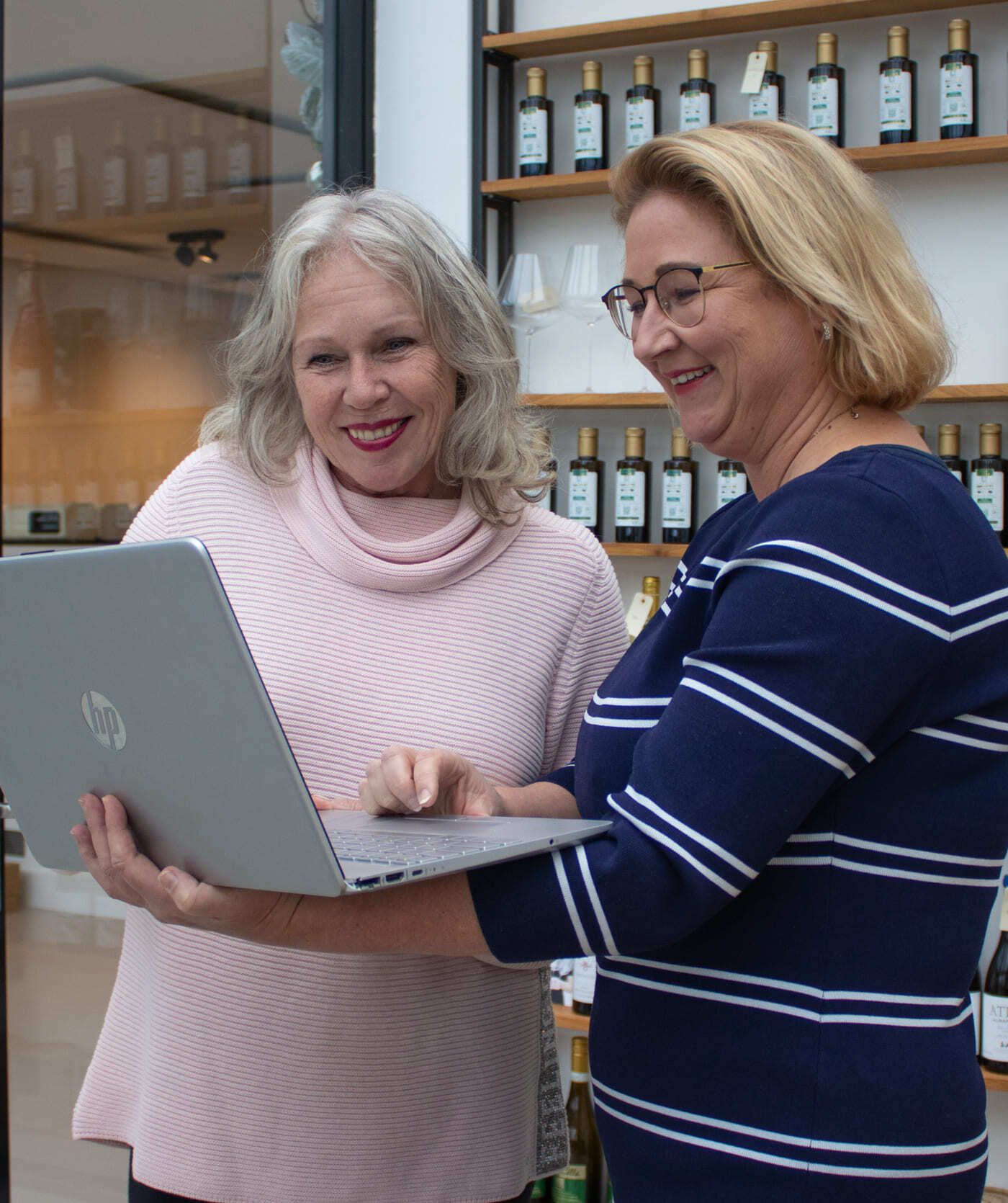 Twee vrouwen staan glimlachend achter een laptop te kijken, mogelijk webshops verkennend. Op de achtergrond zijn schappen met flessen te zien.
