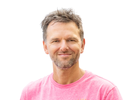 Een man met kort, warrig haar en een baard draagt een roze t-shirt en lacht naar de camera tegen een witte achtergrond. Hij lijkt bereid je te helpen met het bouwen van websites of webshops met professionele online diensten.